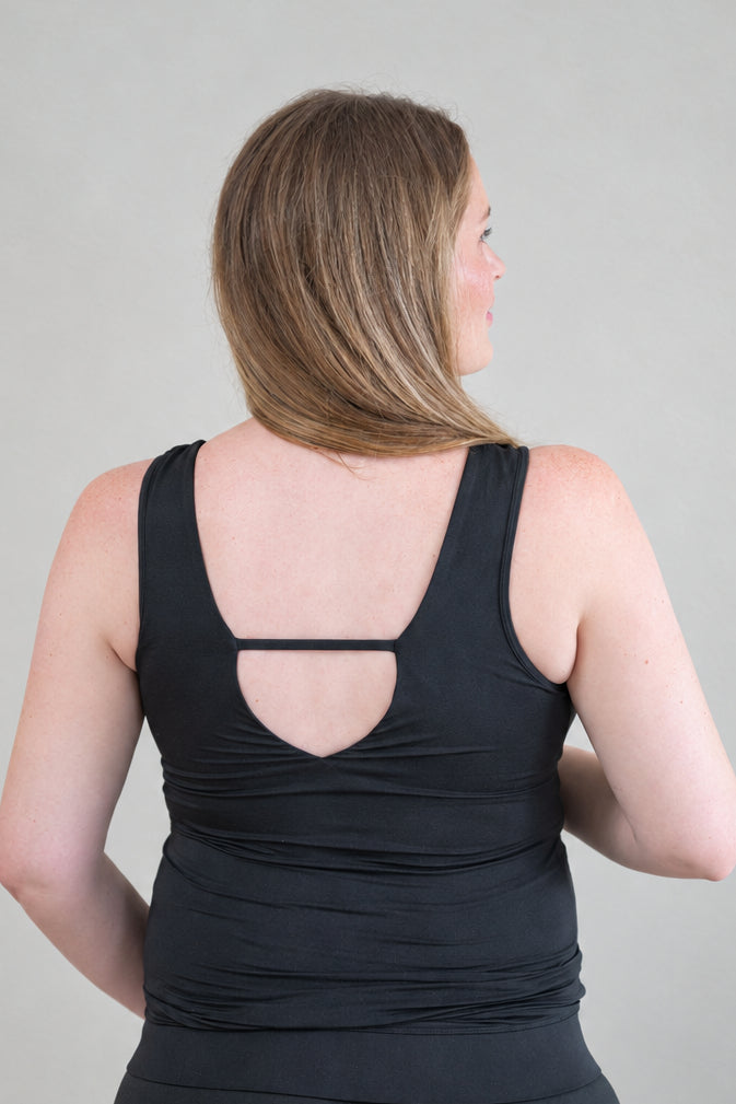 A pregnant woman viewed from the back wearing a black sleeveless top with a cutout detail at the upper back, against a plain light gray studio background.