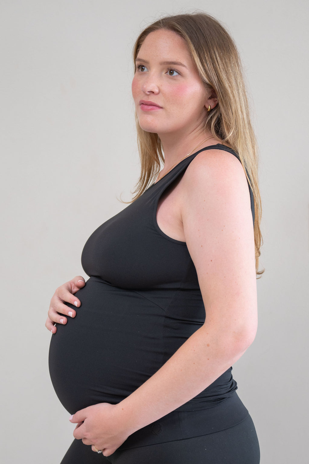 A pregnant woman in a black sleeveless maternity tank top and matching black pants, standing in profile against a light gray studio background.