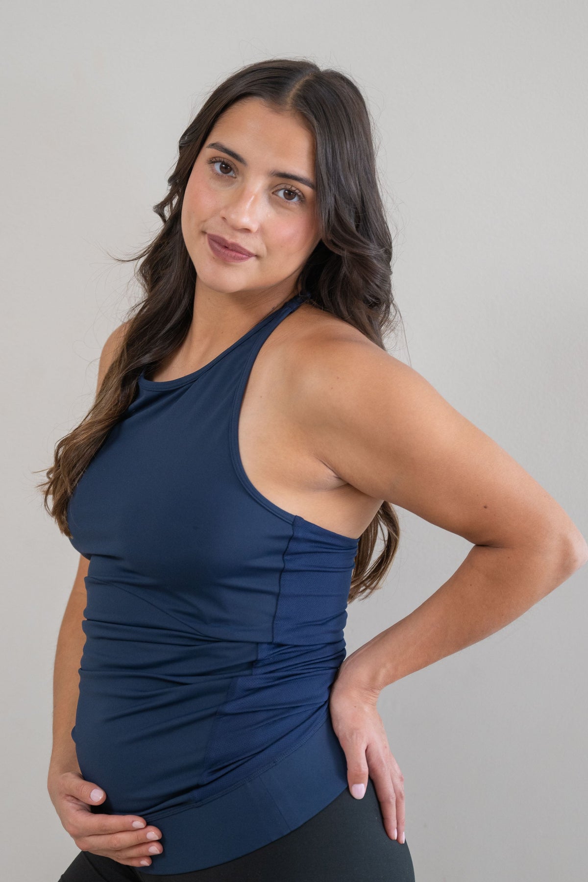 A pregnant woman posing in a navy blue athletic top with braided back and black bike shorts against a plain light gray wall.