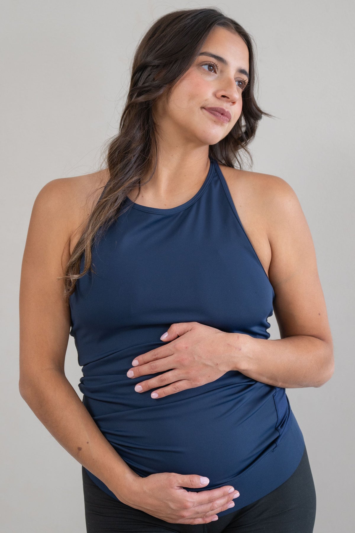 A pregnant woman wearing a navy blue maternity braided back tank and black biker shorts, standing against a neutral light gray background.