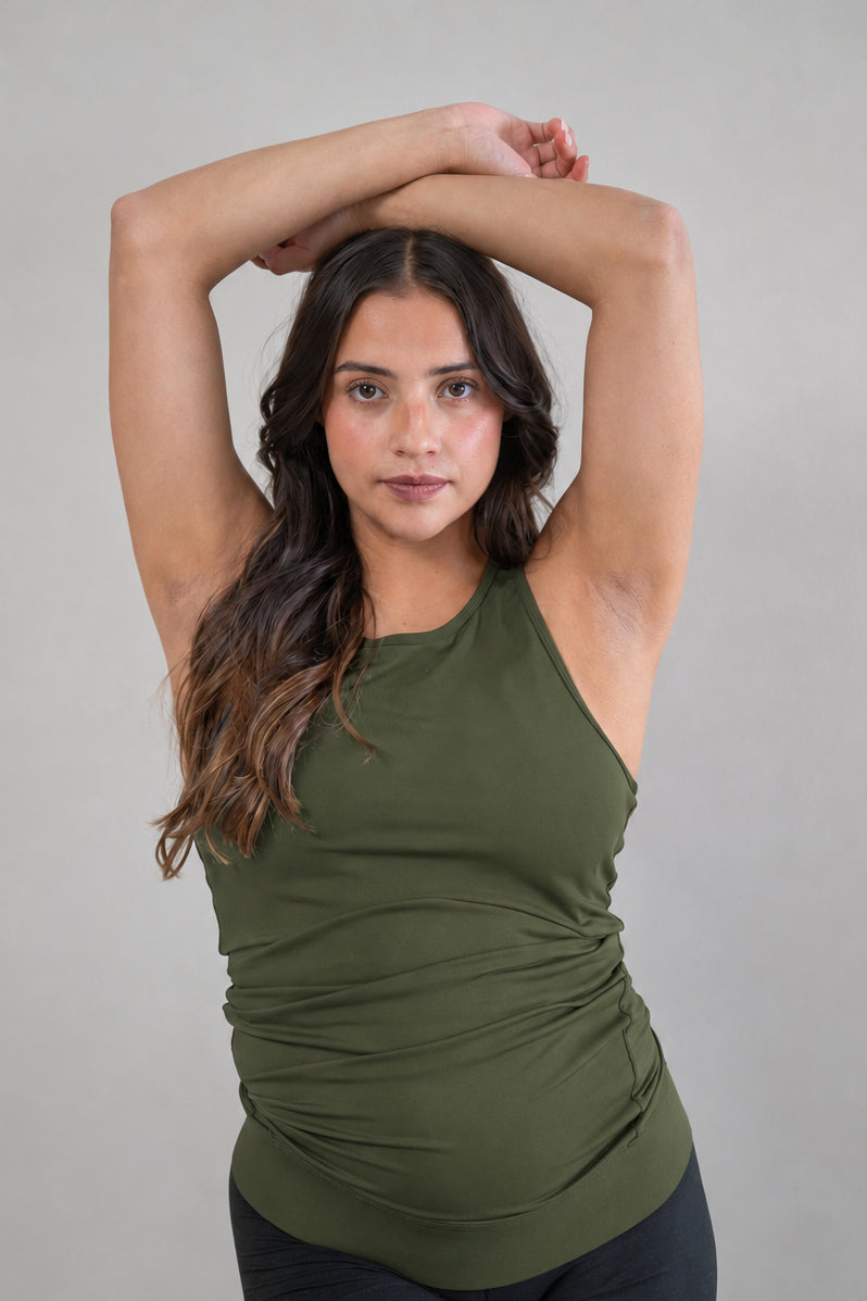 A pregnant woman with long brown hair poses in a green ruched tank top and black bike shorts against a solid light gray background.