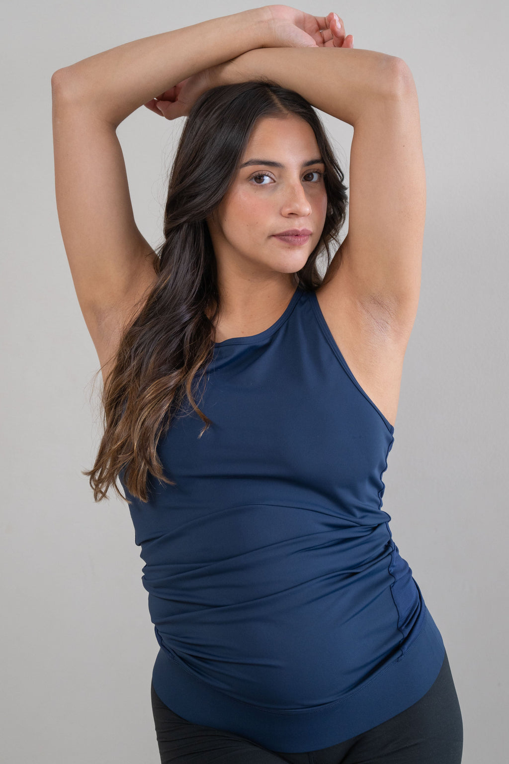 A pregnant woman with long dark brown hair poses in a studio wearing a navy blue ruched halter-neck athletic tank top and black athletic shorts against a solid light gray background.