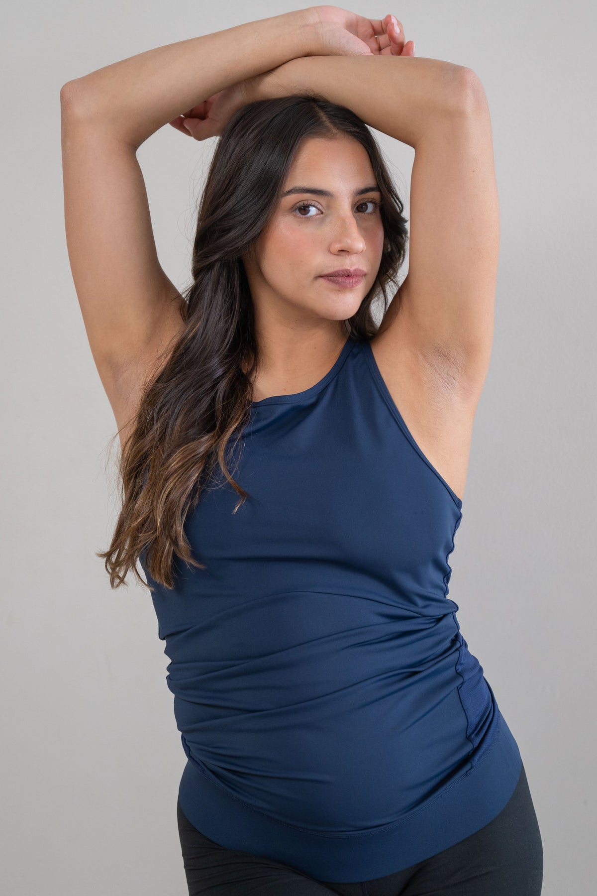A pregnant woman with long dark brown hair poses in a studio wearing a navy blue ruched halter-neck athletic tank top and black athletic shorts against a solid light gray background.