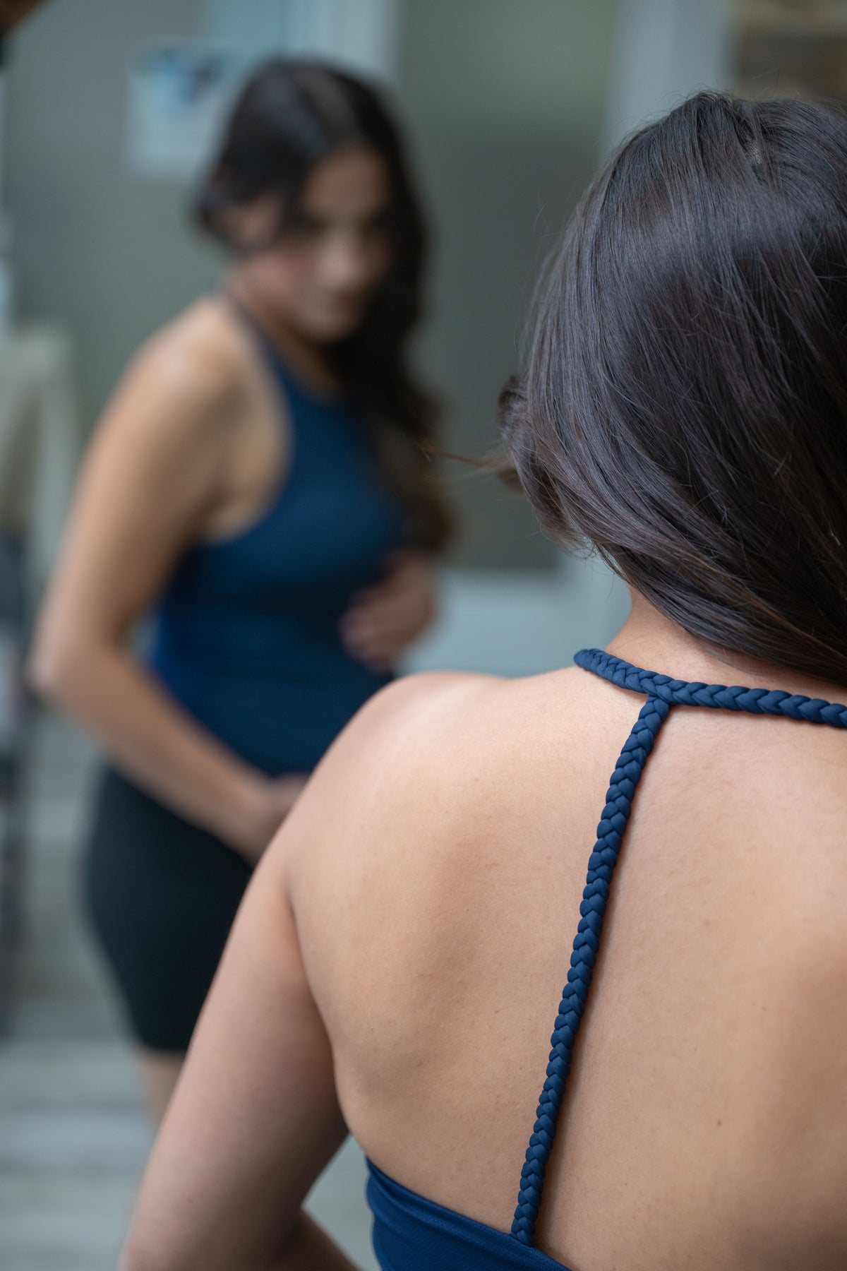 A pregnant woman viewed from behind wearing a navy blue tank with braided back straps, standing in front of a mirror that shows her reflection.