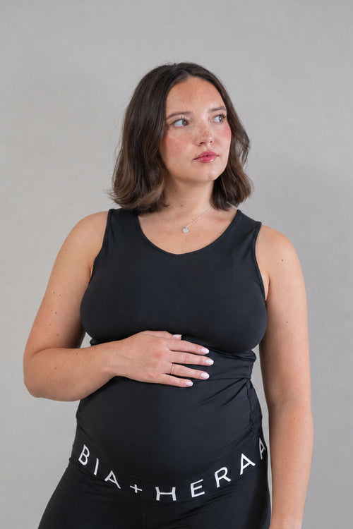 A pregnant woman wearing a black maternity tank top and matching black leggings with the brand name 'BIA+HERA' printed in white on the waistband, standing against a neutral gray background.