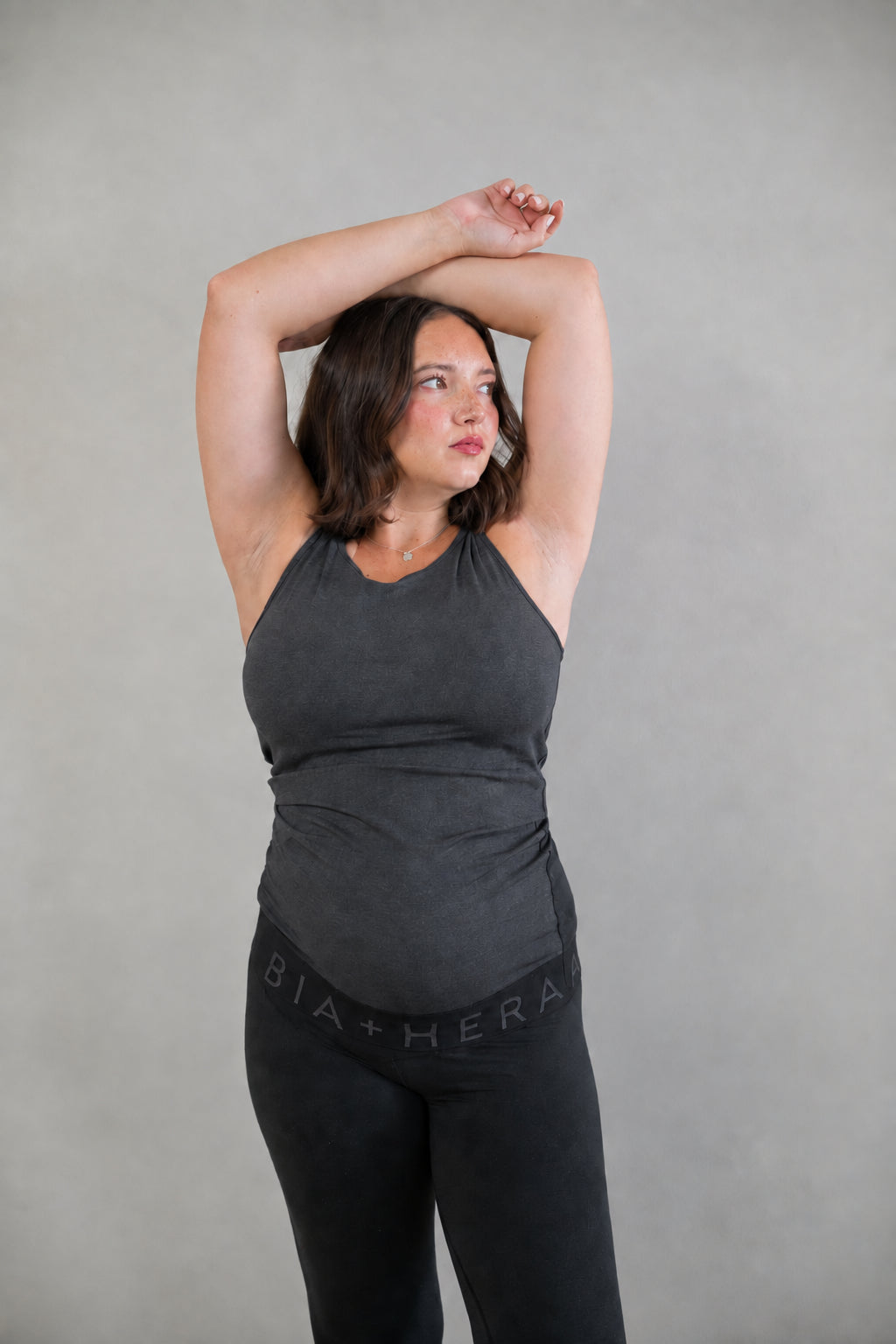 A pregnant woman wearing a dark gray halter tank top and black leggings with 'BIA+HERA' printed on the waistband, posing with her arms raised against a neutral gray background.