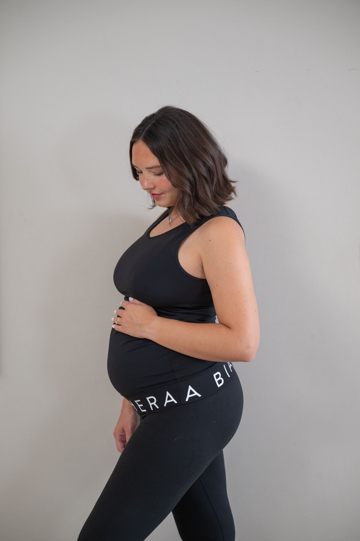 A pregnant woman in a black maternity tank top and black leggings with white text, standing in profile against a plain light gray wall.