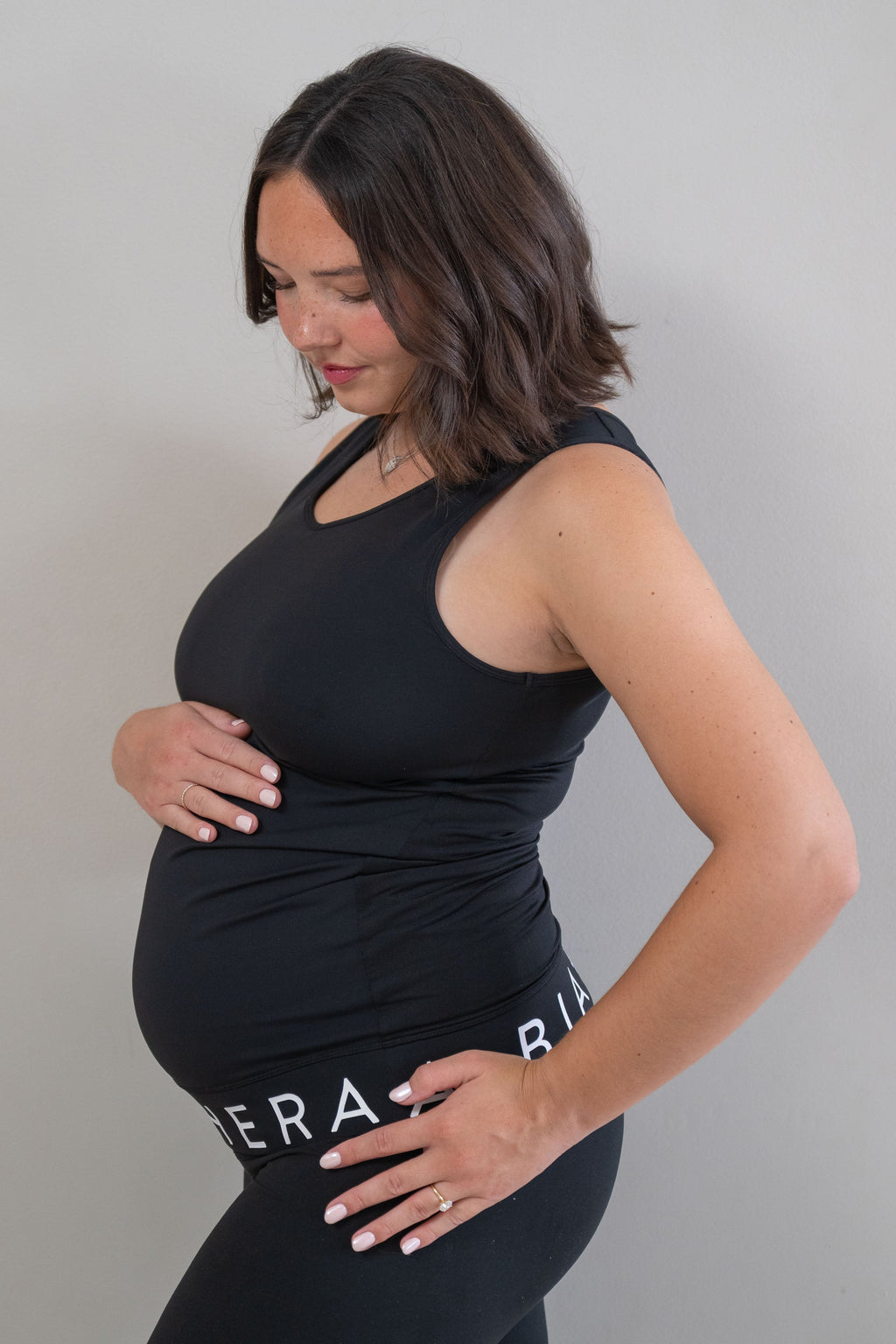A pregnant woman wearing a black maternity tank top and matching black leggings with white text, posing with her hands on her belly against a plain gray background.