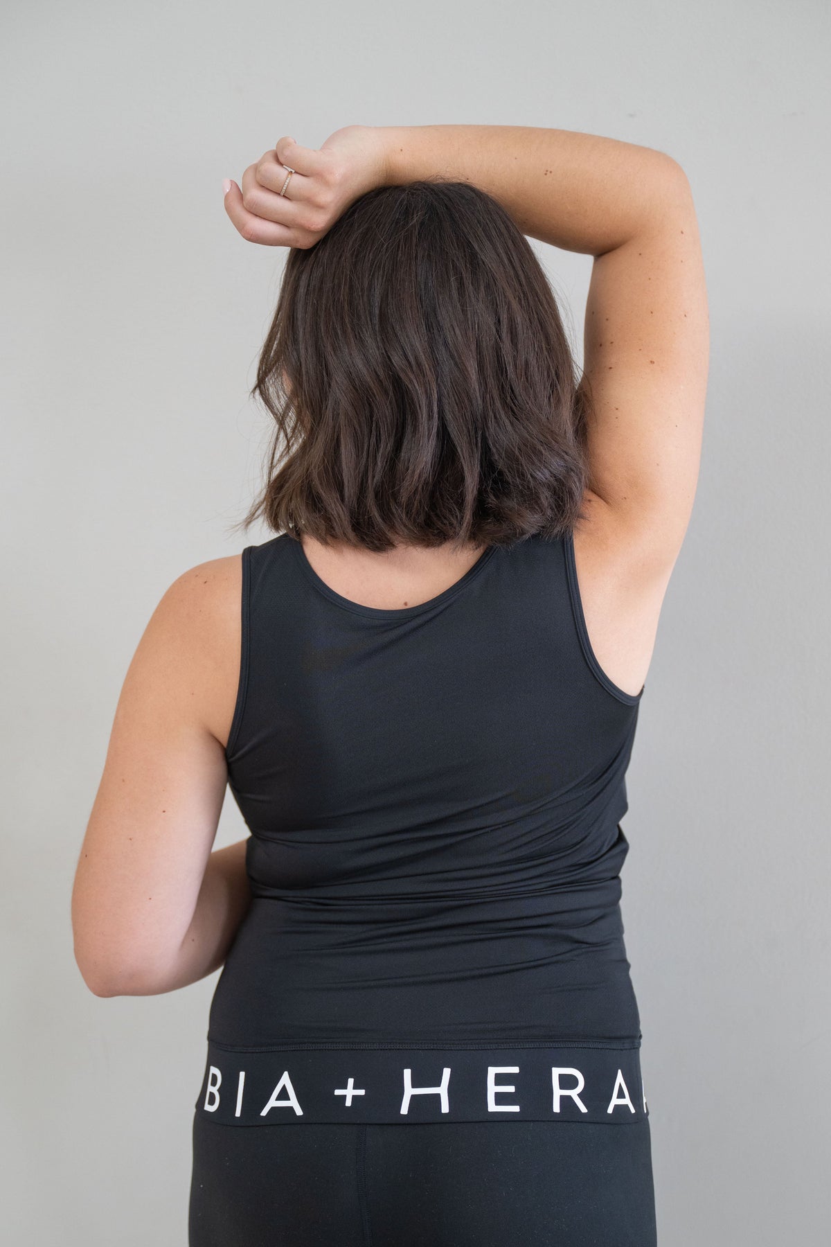 A pregnant woman viewed from behind wearing a black athletic tank top and matching leggings with white text reading 'BIA + HERA' around the waistband, against a plain light gray background.
