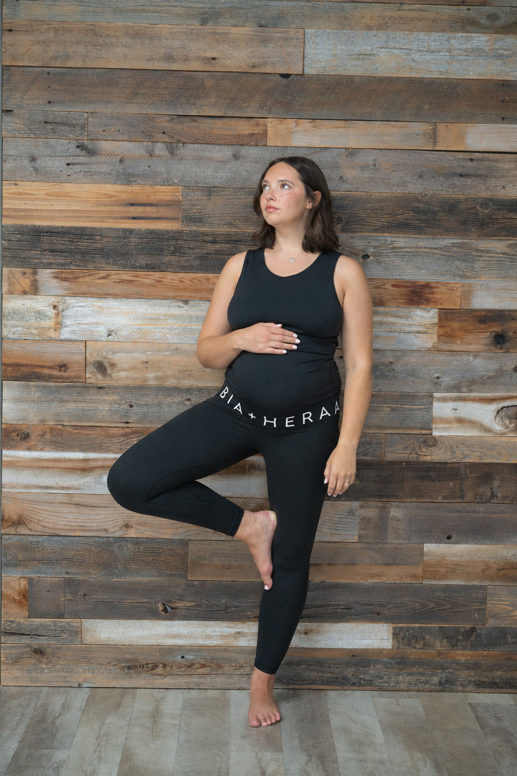 A pregnant woman in a black maternity tank top and black leggings with 'BIA-HERA' printed in white on the hip, posing in a tree pose against a rustic wooden plank wall.