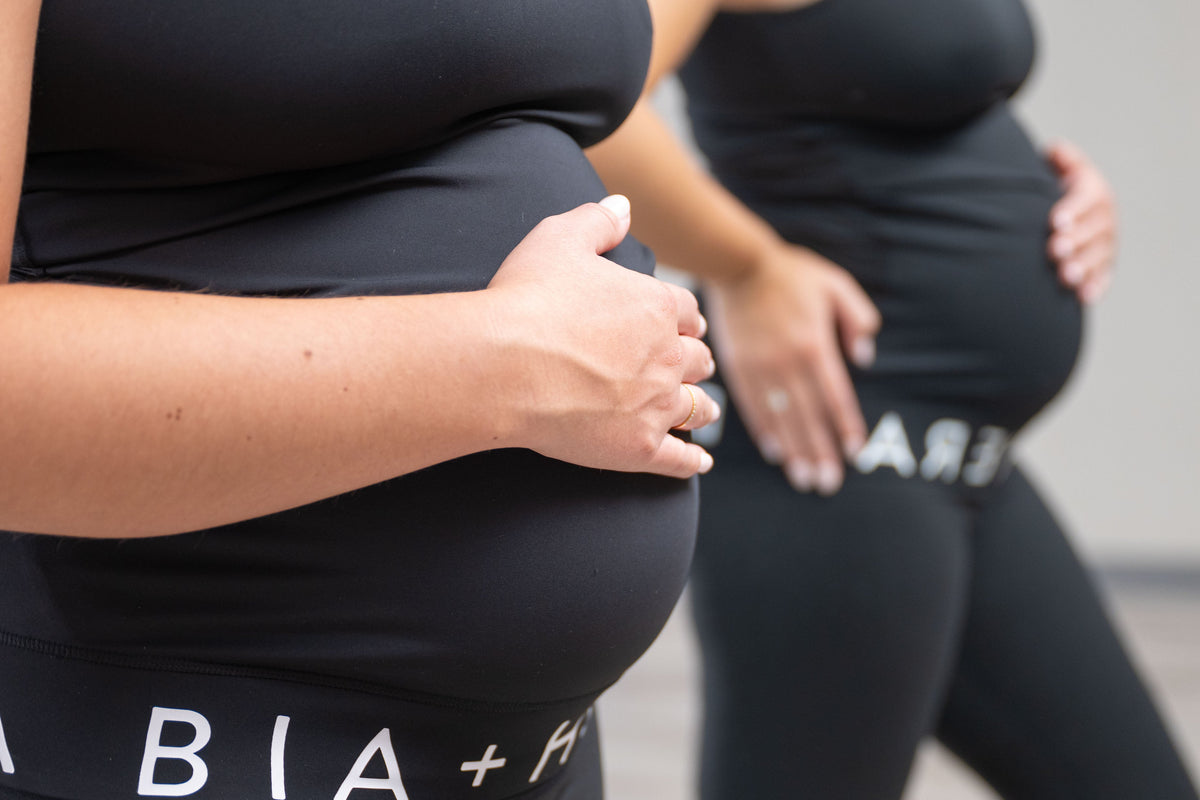 Close-up of a pregnant woman in a black maternity tank top and leggings, with her hands resting on her belly. Another woman is blurred in the background wearing matching black apparel.